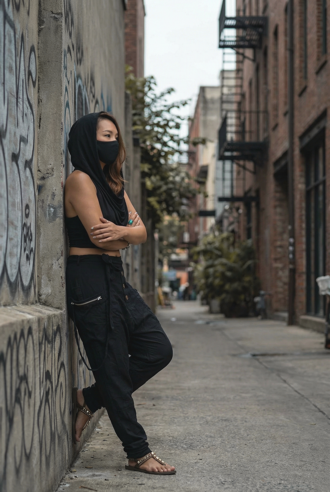 Person wearing a black outfit and mask leaning against a wall in an urban alley.