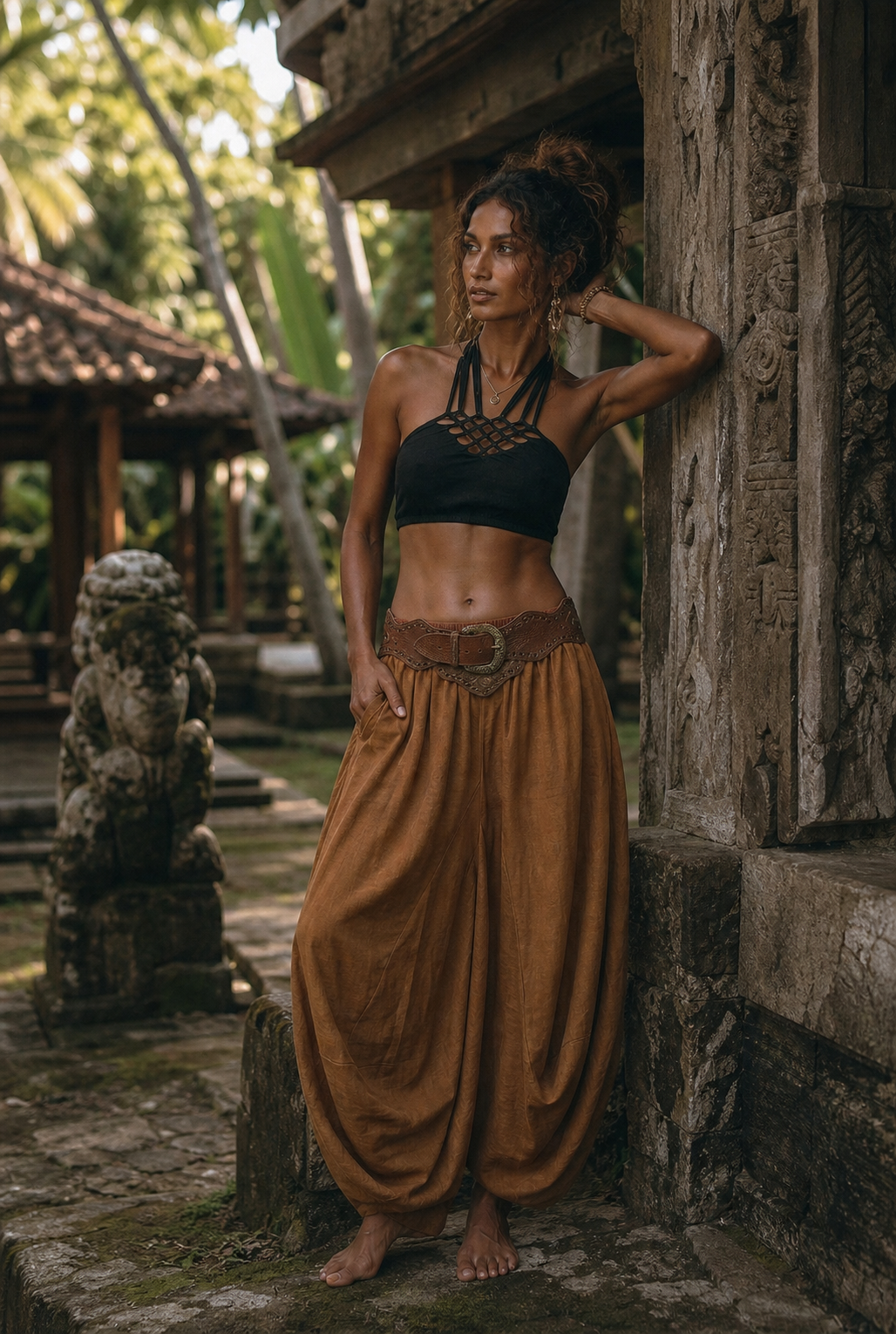 A woman posing outdoors in a tropical setting, wearing a black halter crop top and a brown wide-leg harem pant set with a matching belt.