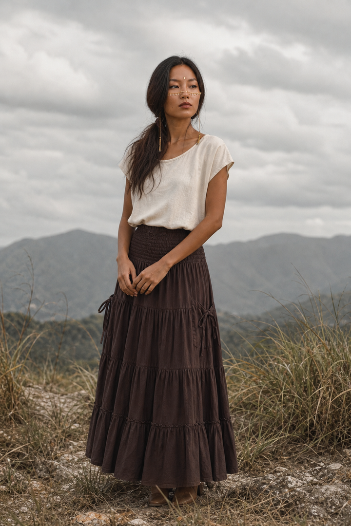 A woman stands outdoors in a grassy field with mountains and an overcast sky in the background, wearing a cream short-sleeve top and a dark brown tiered maxi skirt.
