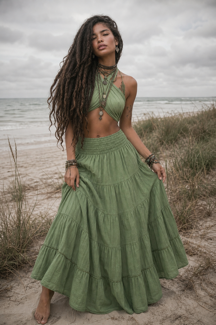 A woman with long dark curly hair stands barefoot on a sandy beach wearing a sage green two-piece set consisting of a halter crop top and a matching tiered maxi skirt. The background shows the ocean, beach grass, and an overcast sky.