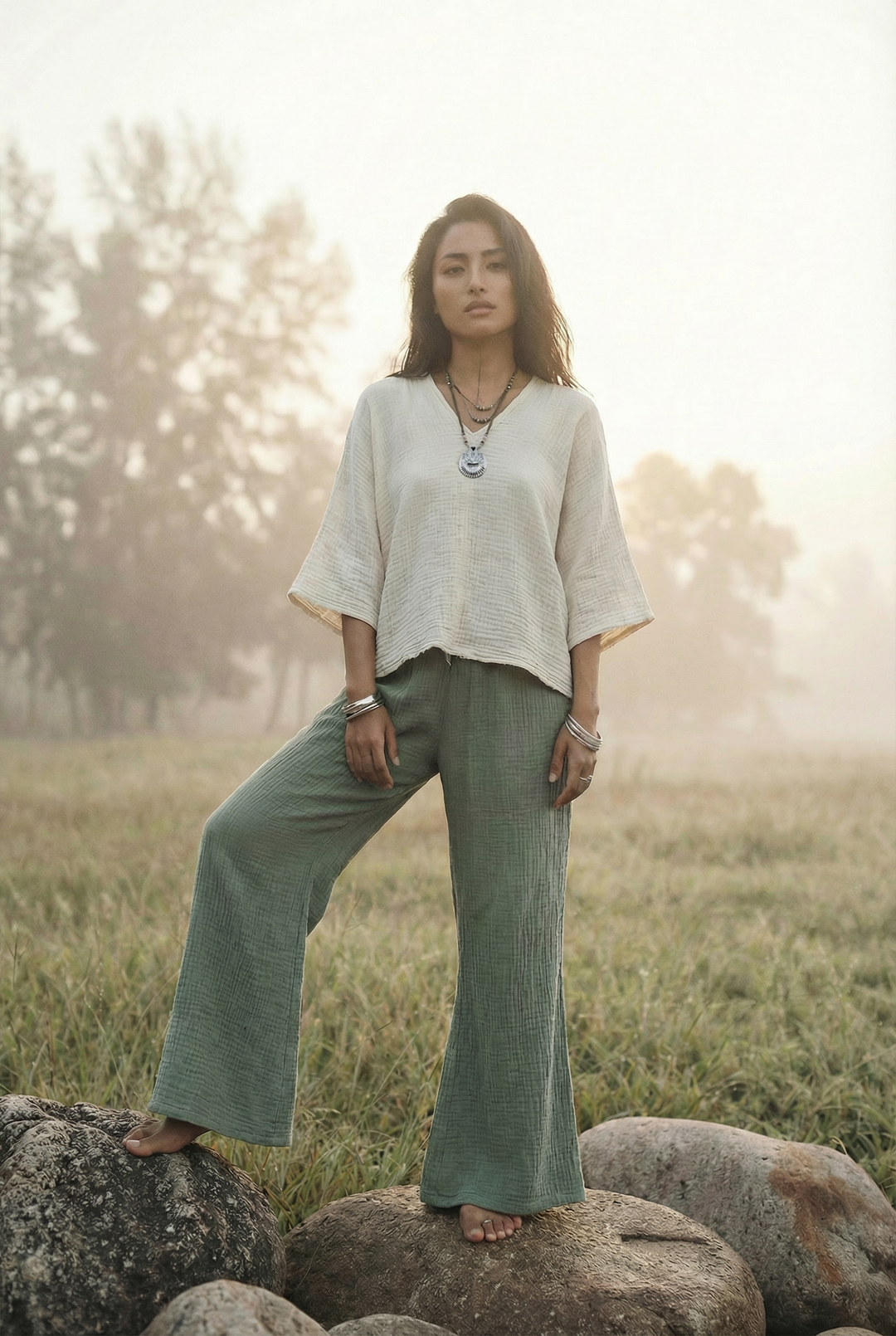 Woman standing on rocks in a field wearing a light blouse and green pants.