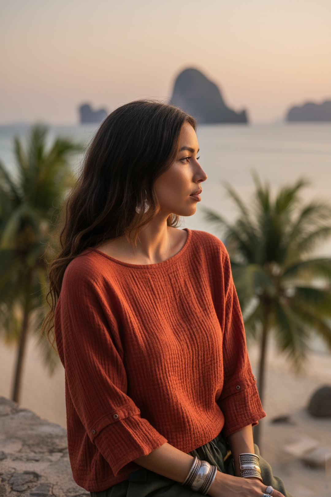 Woman in a rust-colored sweater standing on a beach with palm trees and a sunset in the background