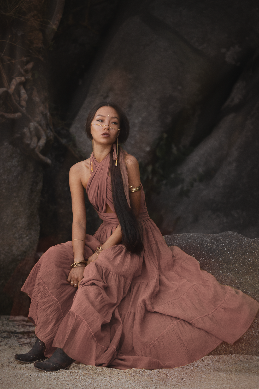 Woman in a long, flowing dress sitting on a rocky surface with a dark, textured background.