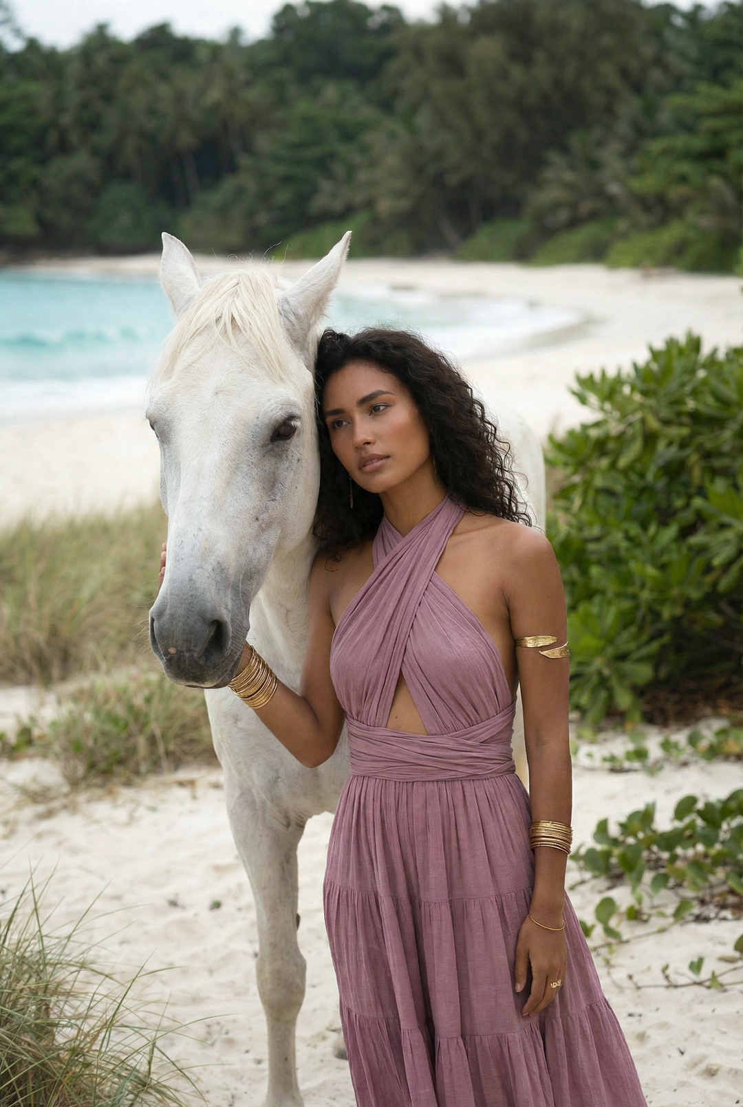 Woman in a pink dress standing next to a white horse on a beach with greenery in the background