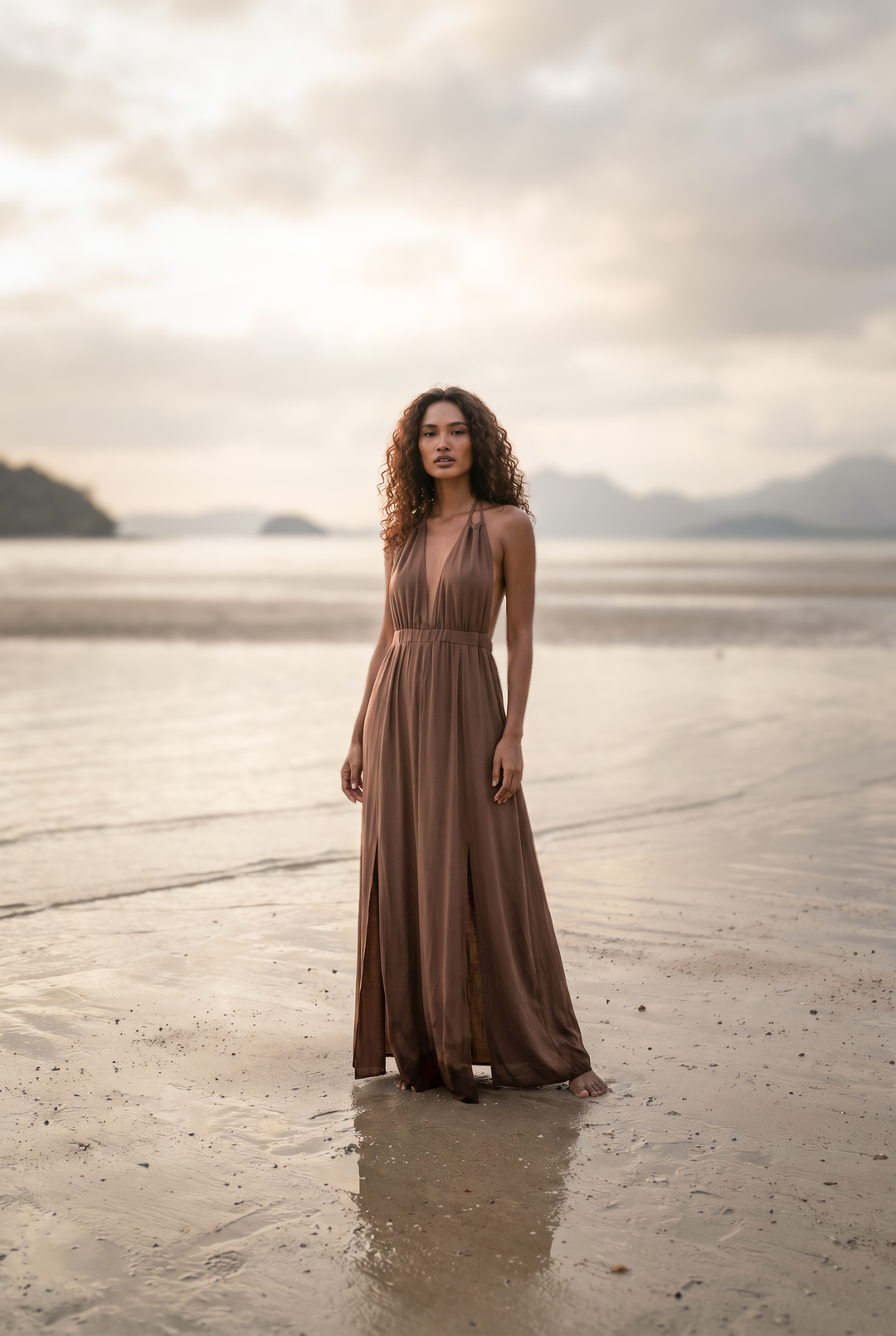 Woman in a long brown dress standing on a beach with mountains in the background