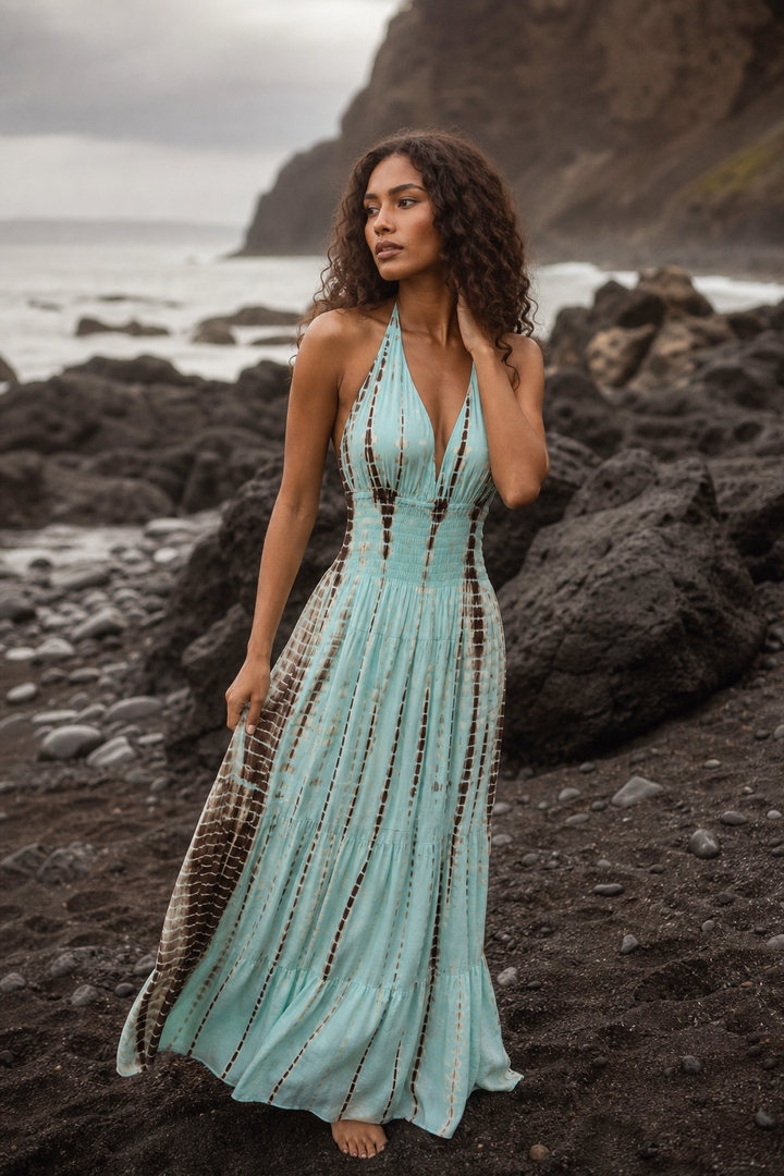 A woman stands on a rocky beach wearing a sleeveless halter maxi dress in light aqua with vertical brown and beige patterned side panels and a deep V-neckline.