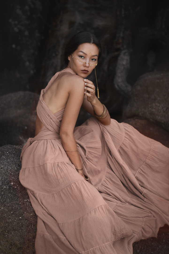 Woman in a long, pink flowing dress sitting on rocks with a dark, natural background