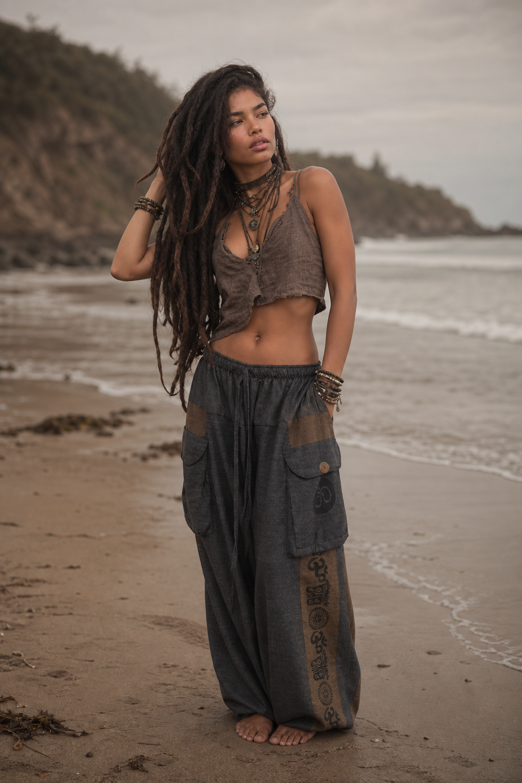 A woman with long dark dreadlocks stands barefoot on a sandy beach wearing a matching two-piece bohemian outfit consisting of a brown cropped tank top and dark gray wide-leg pants with decorative prints.