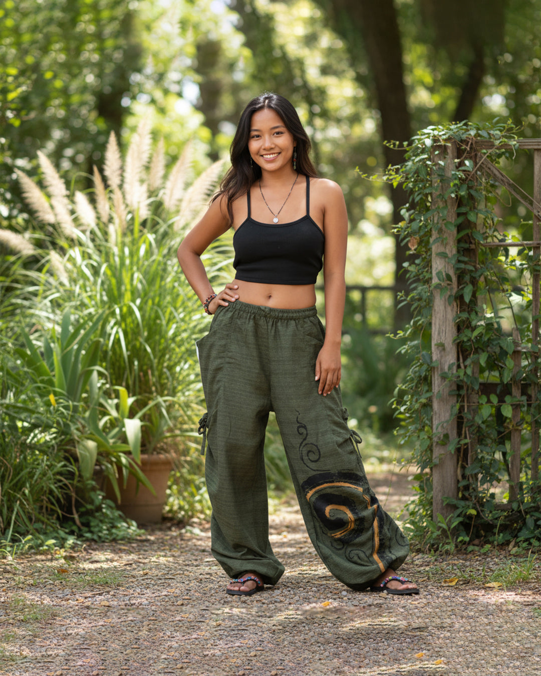 Woman wearing a black top and green baggy pants standing in a garden.