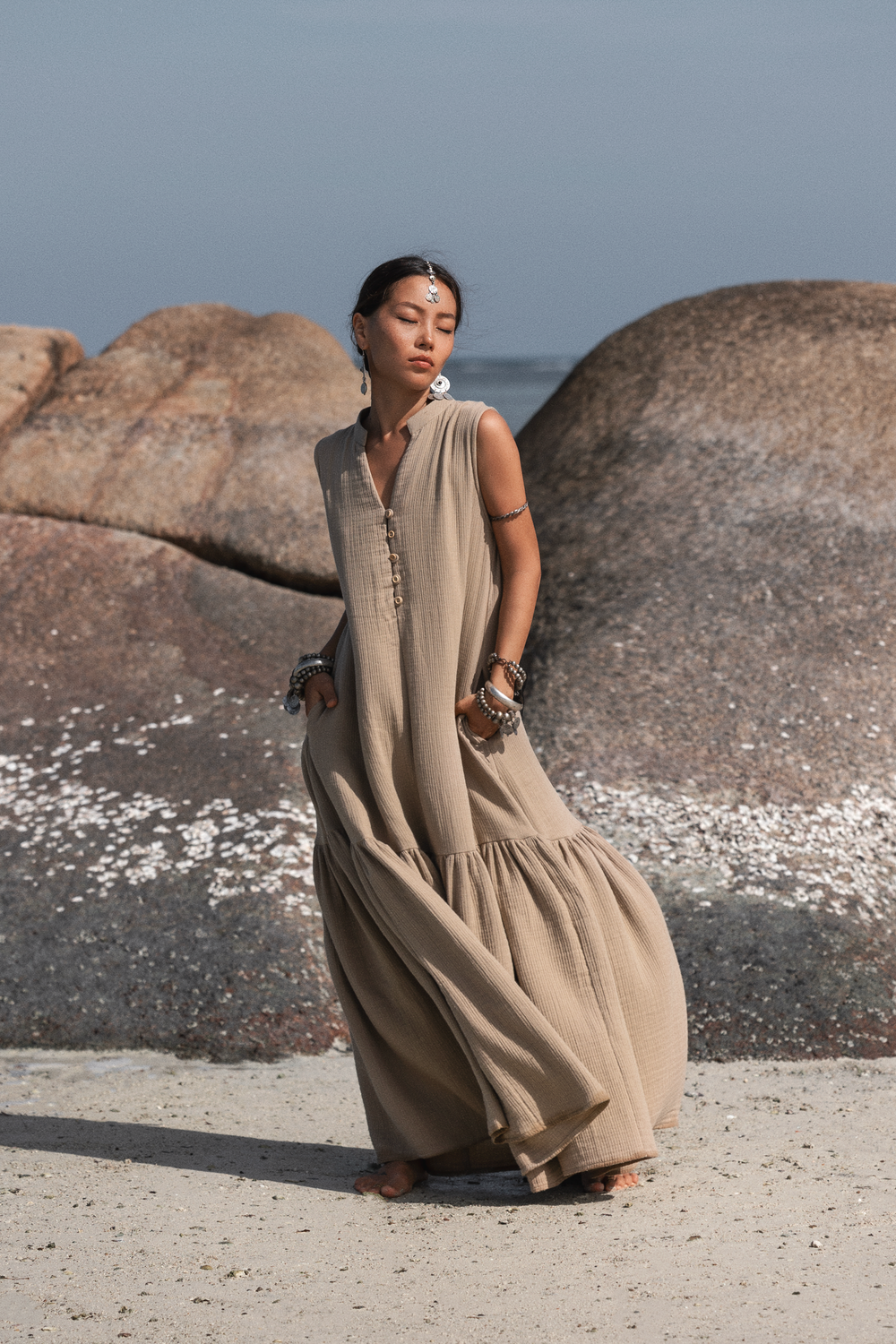 Woman in a long beige dress standing on a rocky beach with ocean in the background