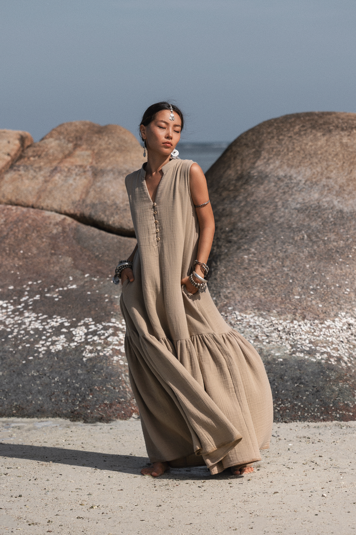 Woman in a long beige dress standing on a rocky beach with ocean in the background