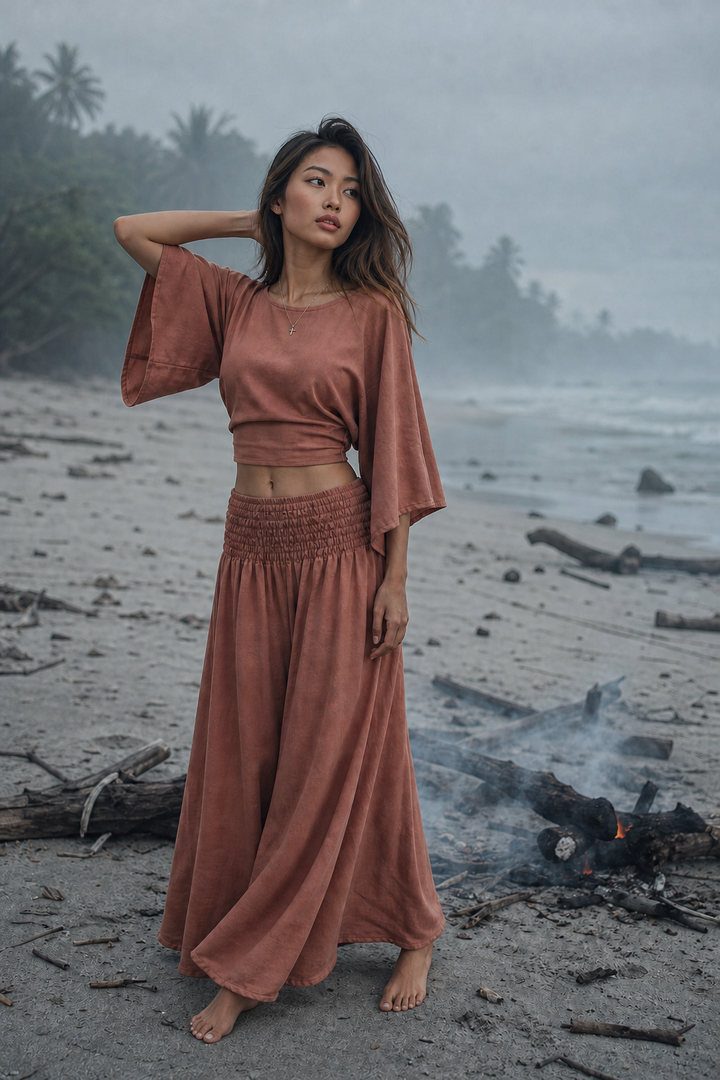 A woman stands barefoot on a beach wearing a matching rust-colored two-piece outfit with a cropped short-sleeve top and a long smocked maxi skirt. The background shows a misty shoreline with palm trees, ocean water, and driftwood.