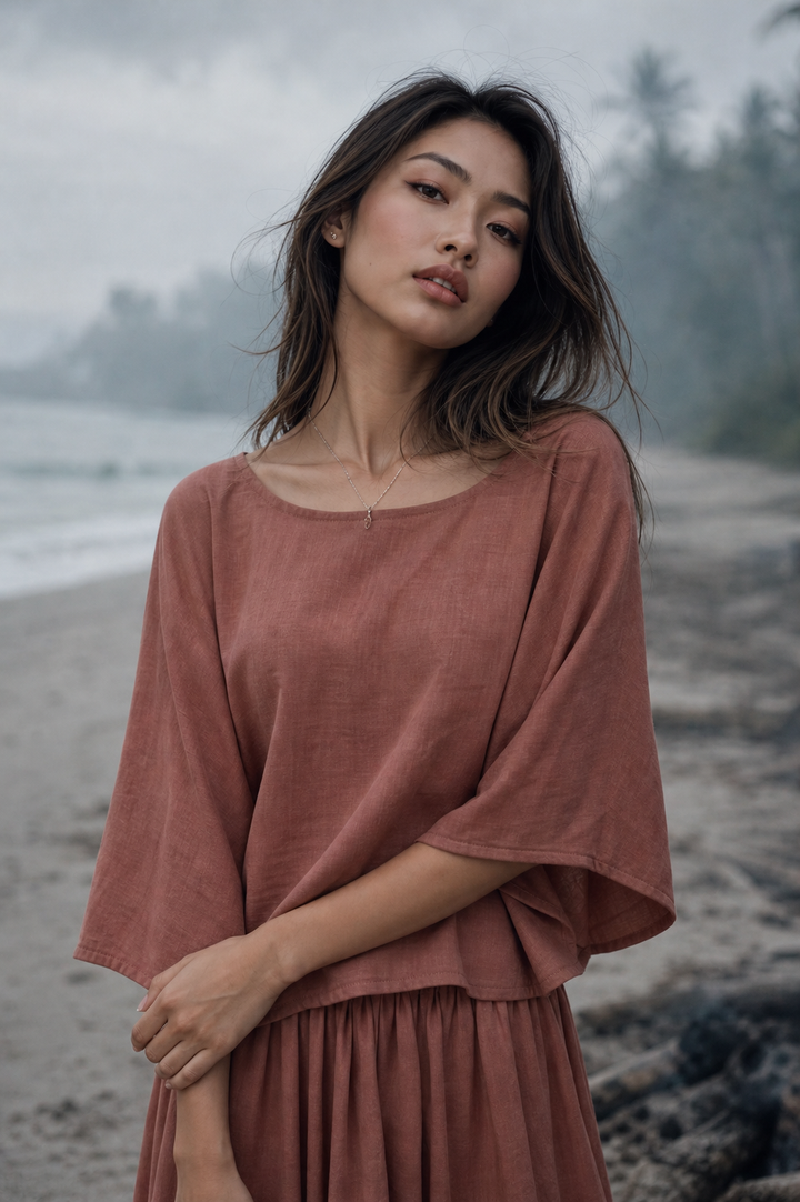 A woman wearing a loose, dusty rose linen dress stands on a beach with a misty, blurred background of water and trees.