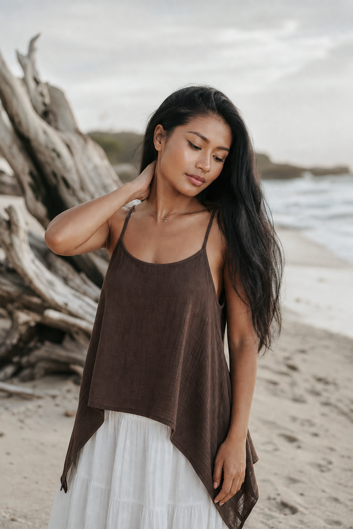 Woman in a brown top and white skirt standing on a beach with driftwood.