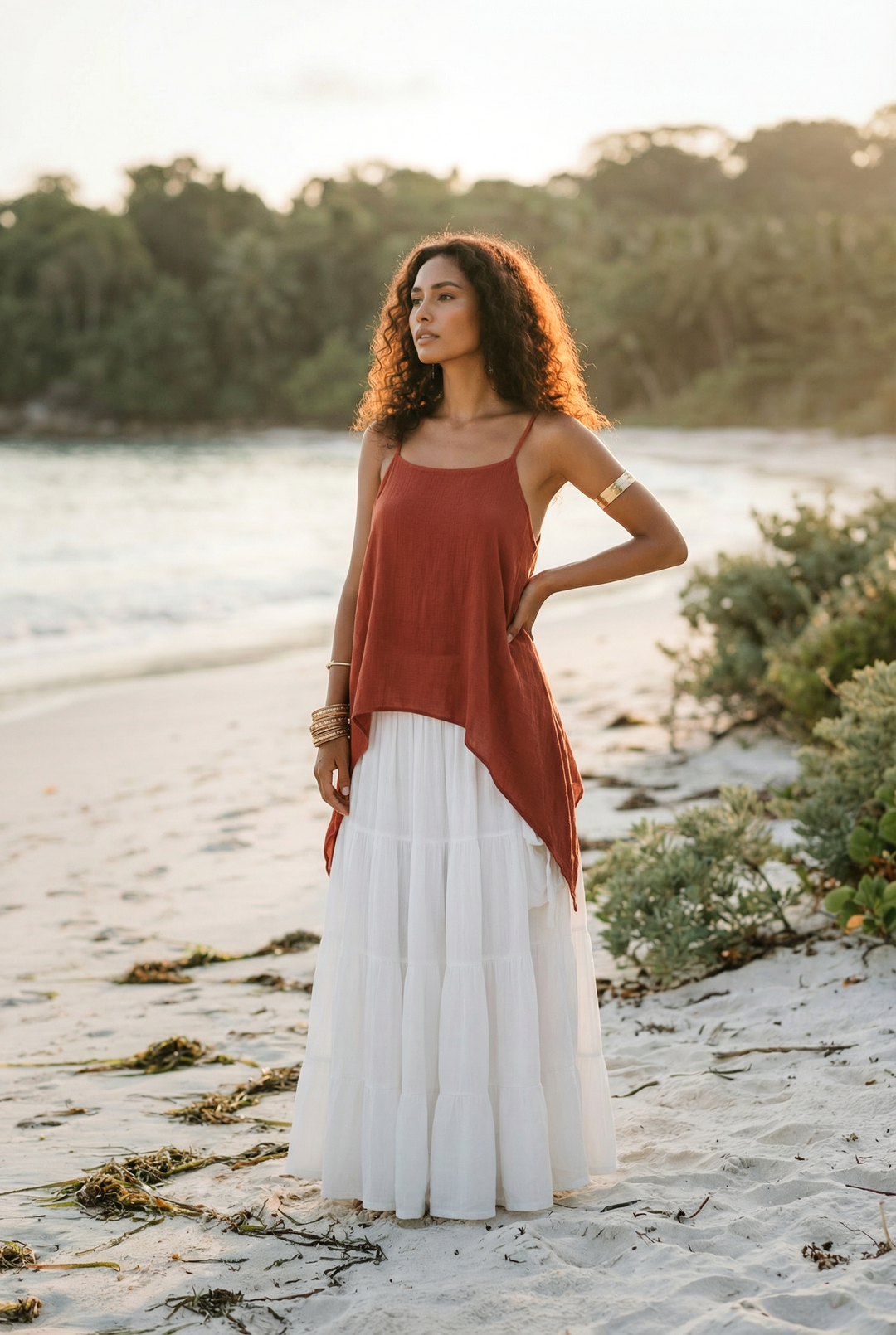 Woman in a rust top and white skirt standing on a beach with trees in the background
