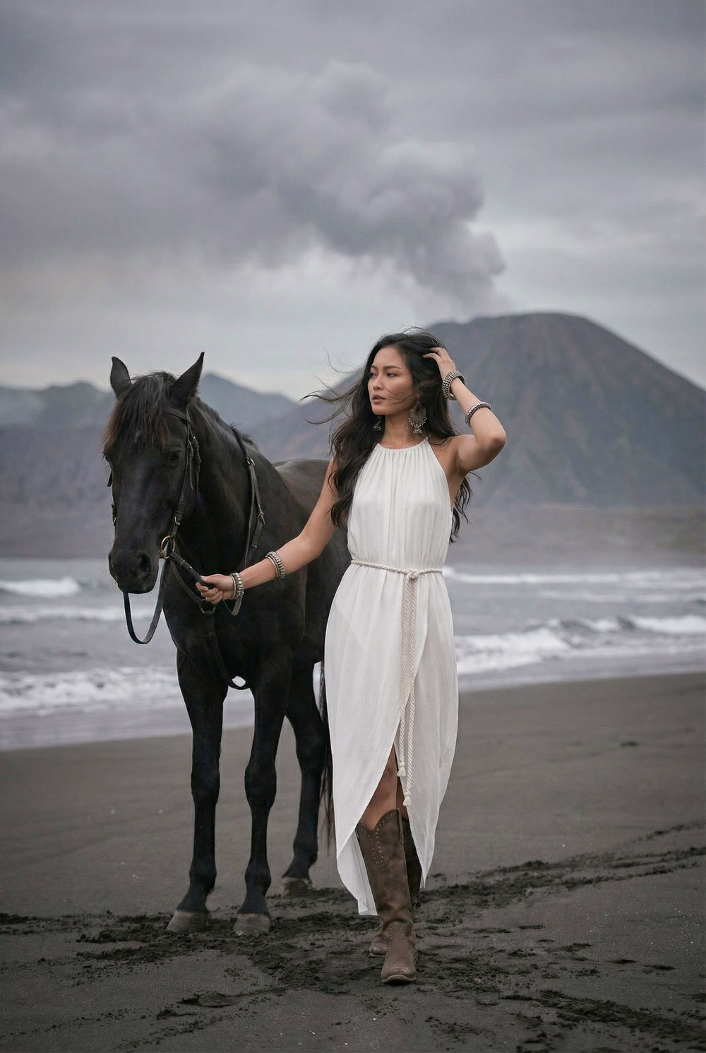 Woman in a white dress standing next to a horse on a beach with a mountain in the background
