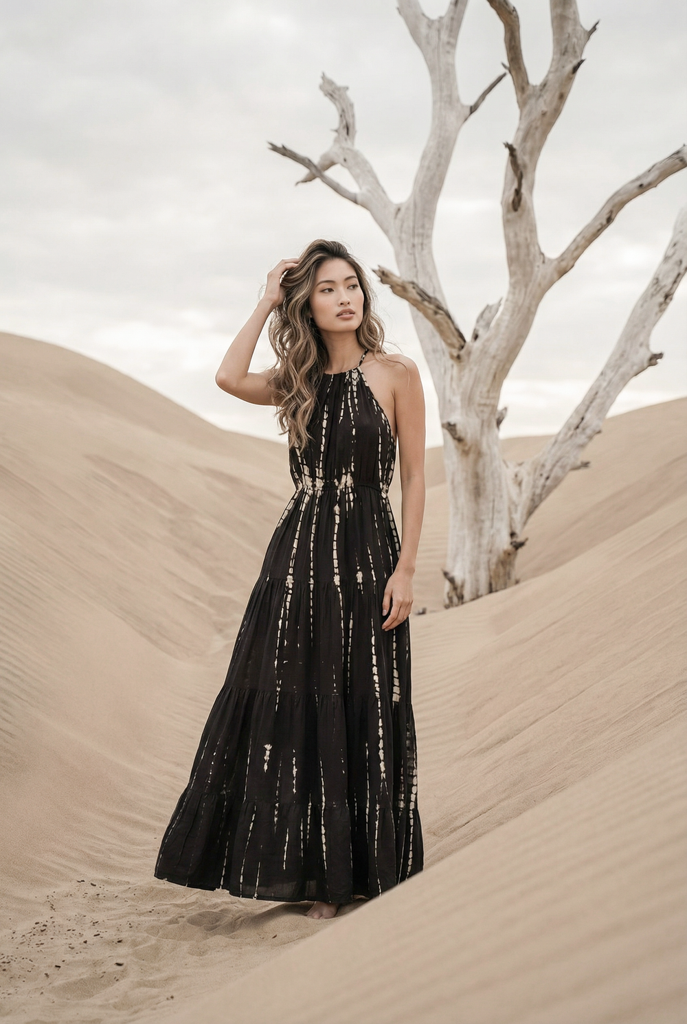 Woman in a black dress standing in sand dunes with a bare tree.