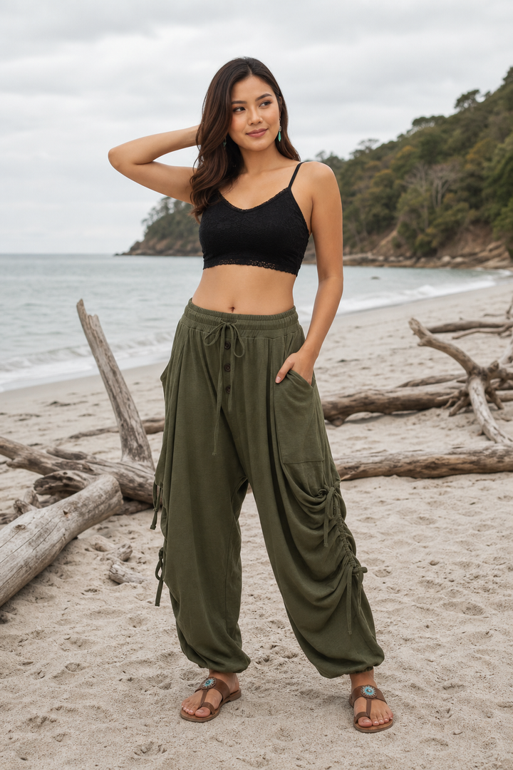 A woman stands on a sandy beach wearing loose olive green drawstring cargo pants and a black crop top, with ocean water, driftwood, and a tree-lined shore in the background.