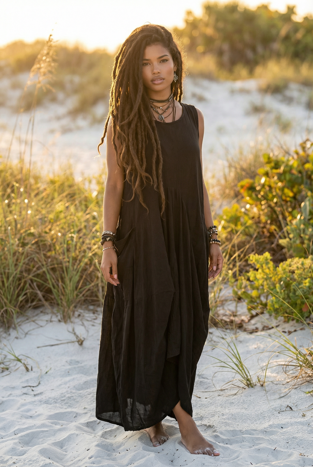 Woman in a black dress standing on a sandy beach with grass and sand dunes in the background.