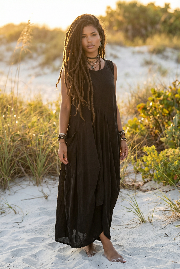 Woman in a black dress standing on a sandy beach with grass and sand dunes in the background.