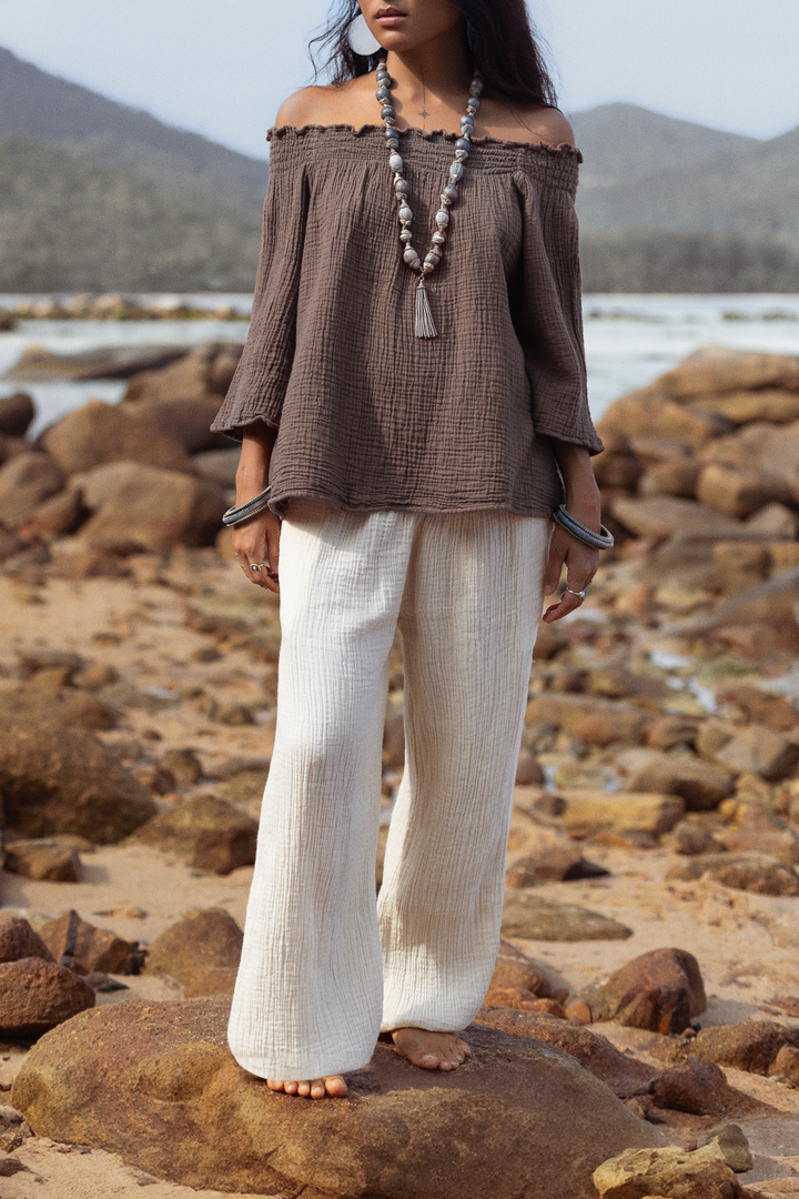 Woman standing on a rocky beach wearing a brown off-shoulder top and white pants.
