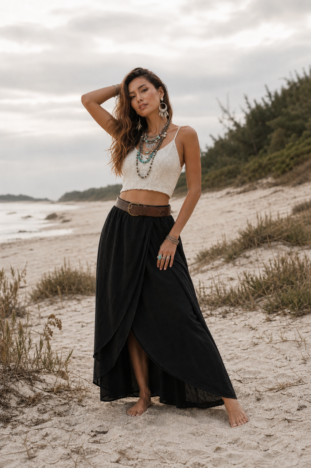 A woman stands barefoot on a sandy beach wearing a white sleeveless crop top and a long black maxi skirt with a brown belt. She is accessorized with layered turquoise necklaces and large hoop earrings, with dunes and water in the background under an overcast sky.
