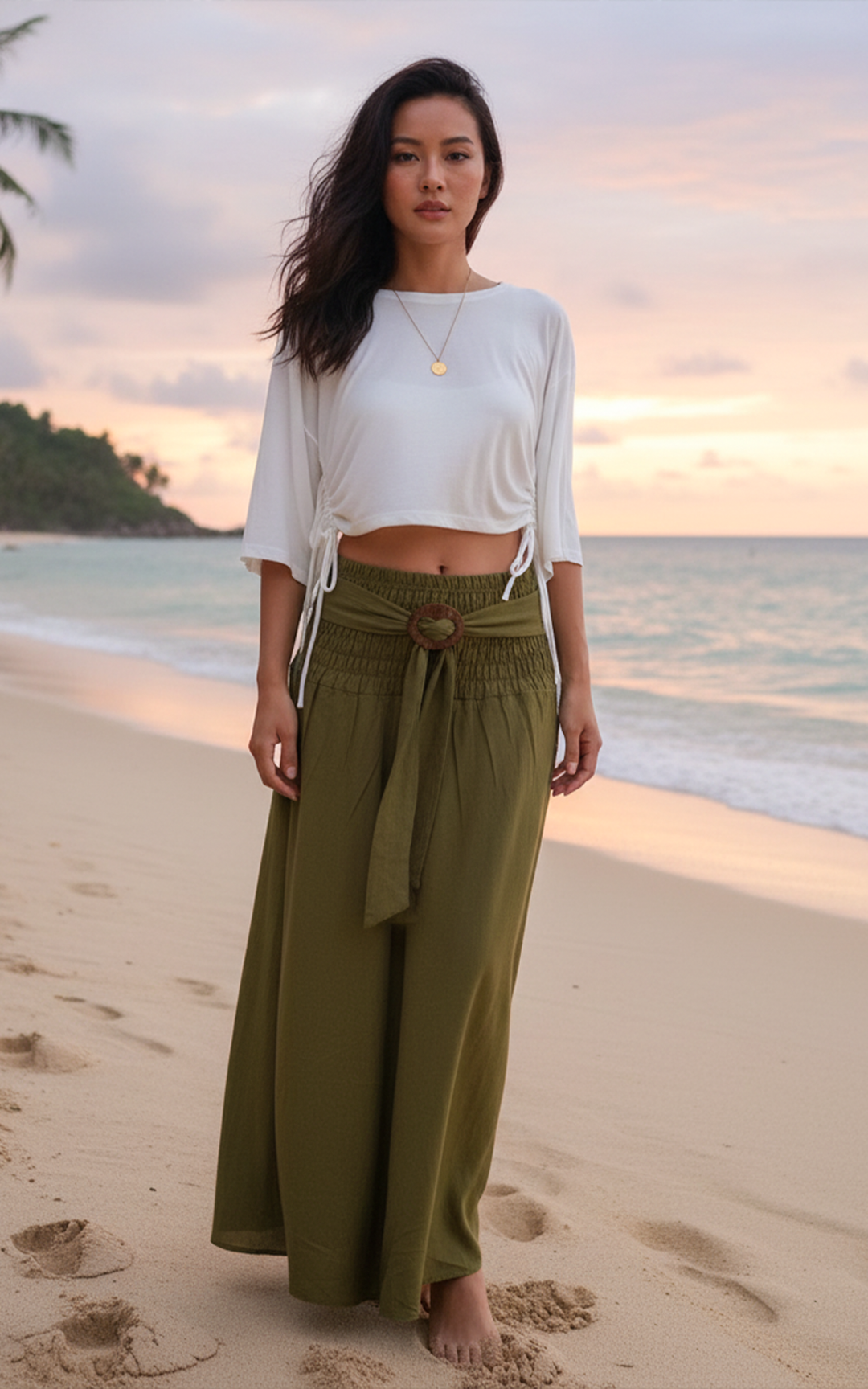 Woman in white top and green pants standing on a beach at sunset.