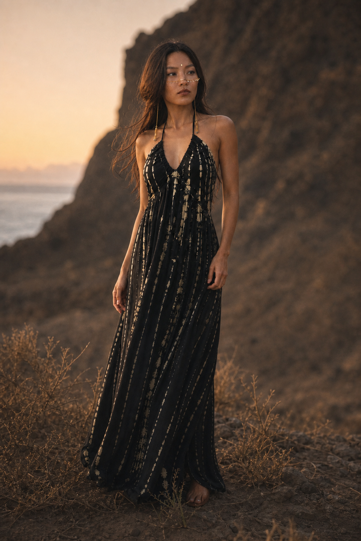 A woman stands on a rocky cliffside outdoors at sunset wearing a long black halter maxi dress with vertical metallic gold stripe patterns. The background features dry grass, a large rock formation, and a soft orange sky with a hint of water on the horizon.