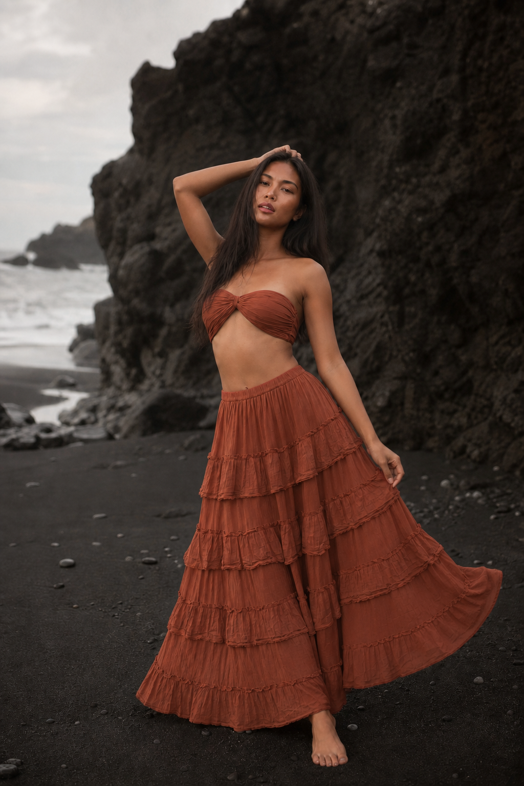 A woman stands barefoot on a black sand beach in front of large dark rock formations, wearing a rust-orange strapless bandeau top and matching tiered maxi skirt.