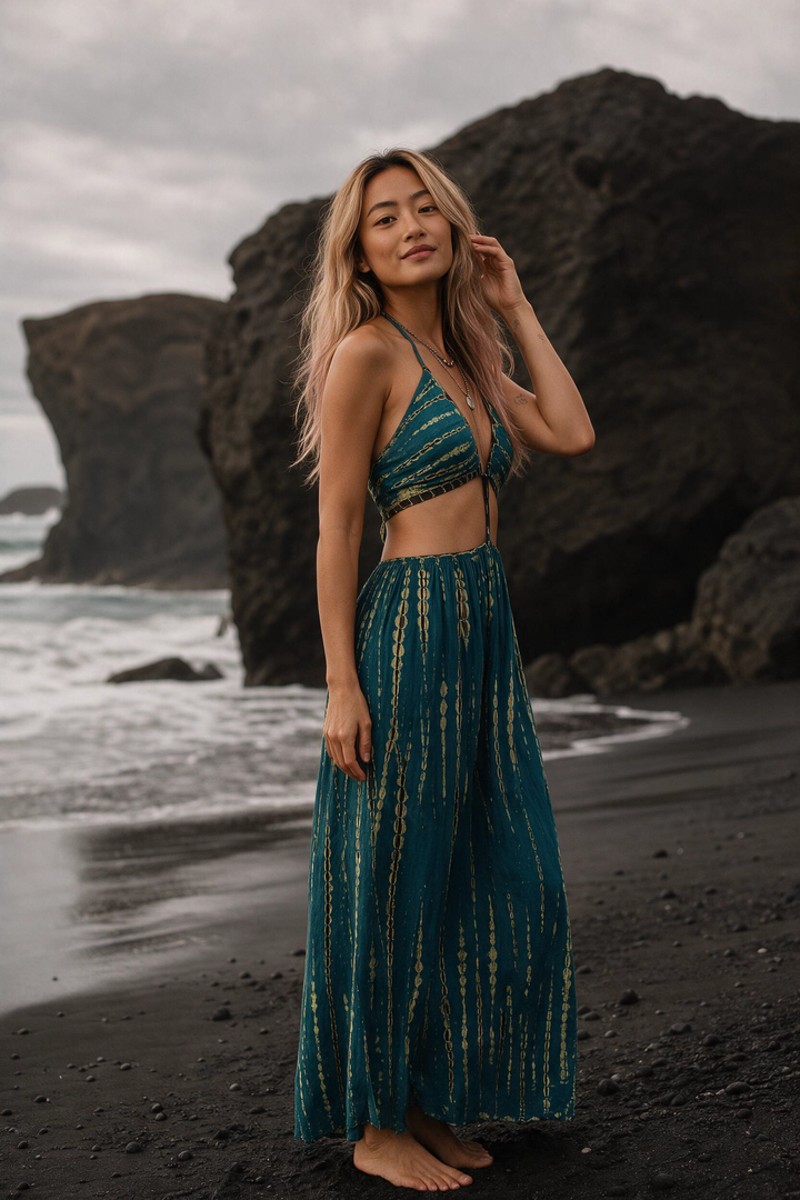 A woman stands barefoot on a black sand beach in front of large dark volcanic rocks, wearing a teal and gold tie-dye two-piece swimsuit consisting of a halter crop top and a matching maxi skirt.