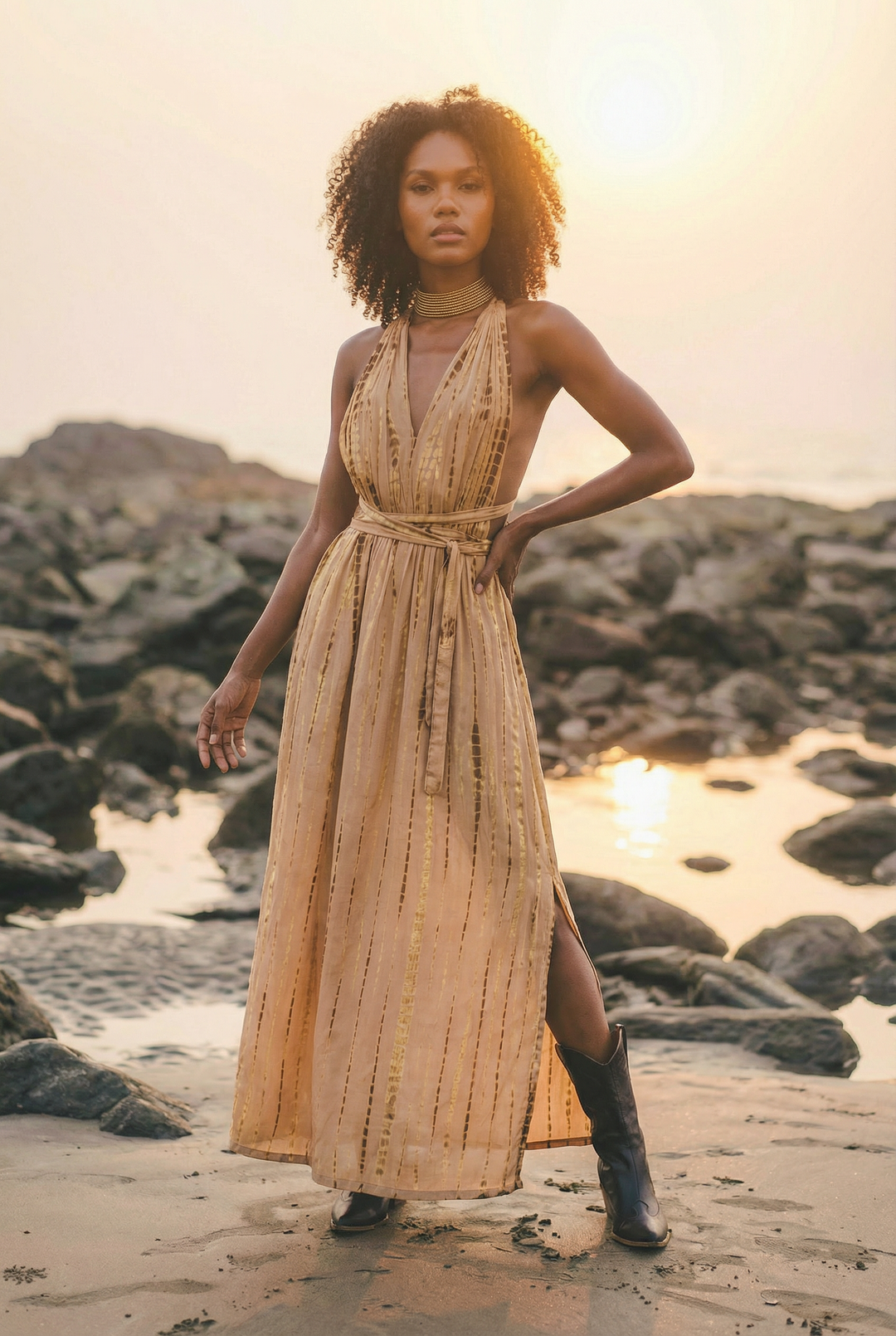 Woman in a long beige dress standing on a rocky beach with sunset in the background