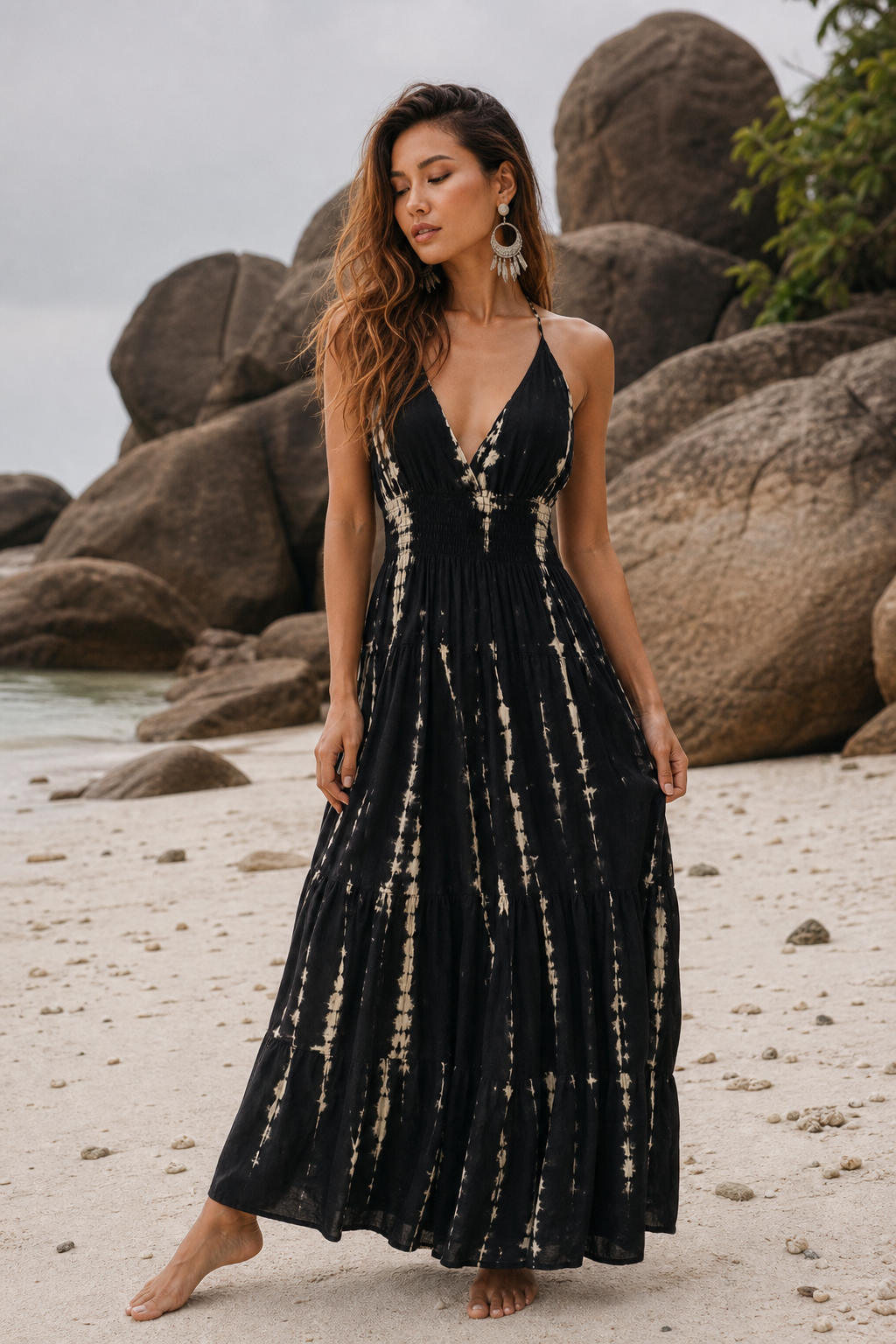 A woman stands barefoot on a sandy beach wearing a sleeveless black maxi dress with a beige tie-dye pattern. Large rock formations and the ocean are visible in the background.