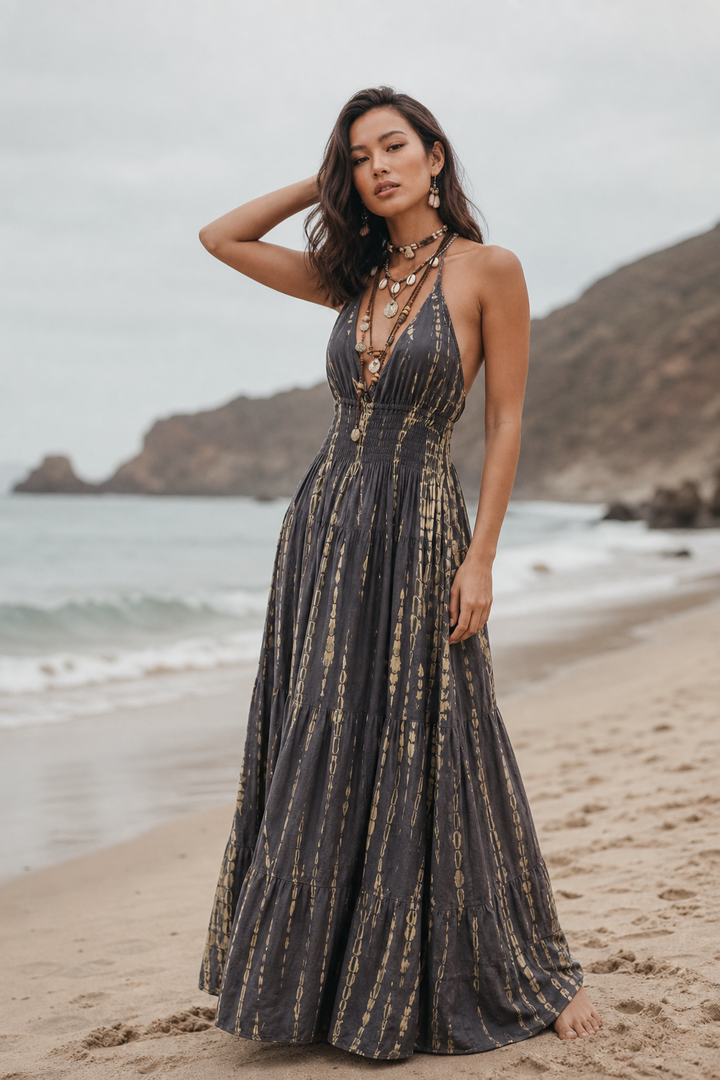 A woman stands on a sandy beach wearing a long dark gray maxi dress with vertical gold-toned metallic stripes. The background features the ocean, distant rocky cliffs, and an overcast sky.