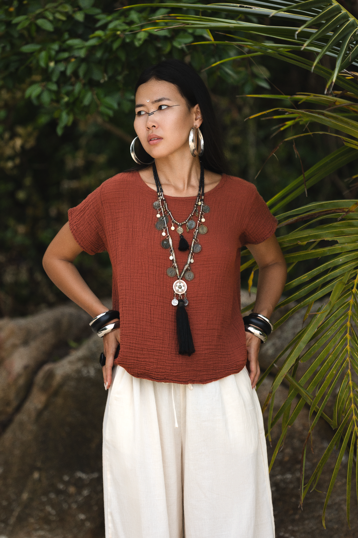 Woman wearing a rust-colored top and white pants with a necklace, standing against a natural background.